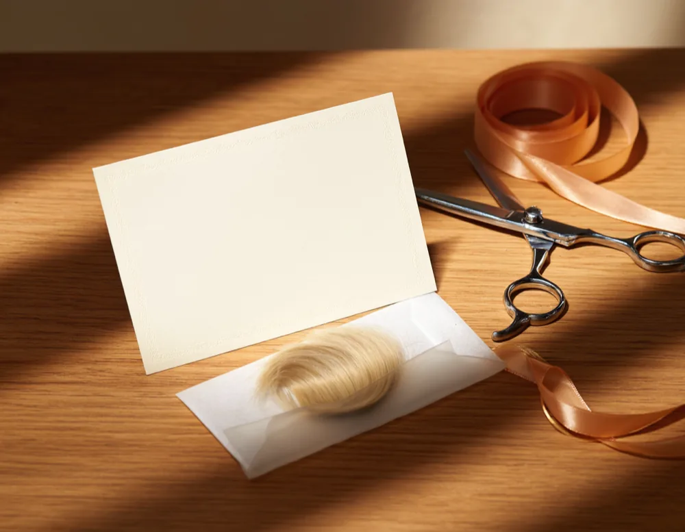 A blank certificate card on a wooden counter next to a glassine envelope holding a lock of light hair, scissors and a ribbon