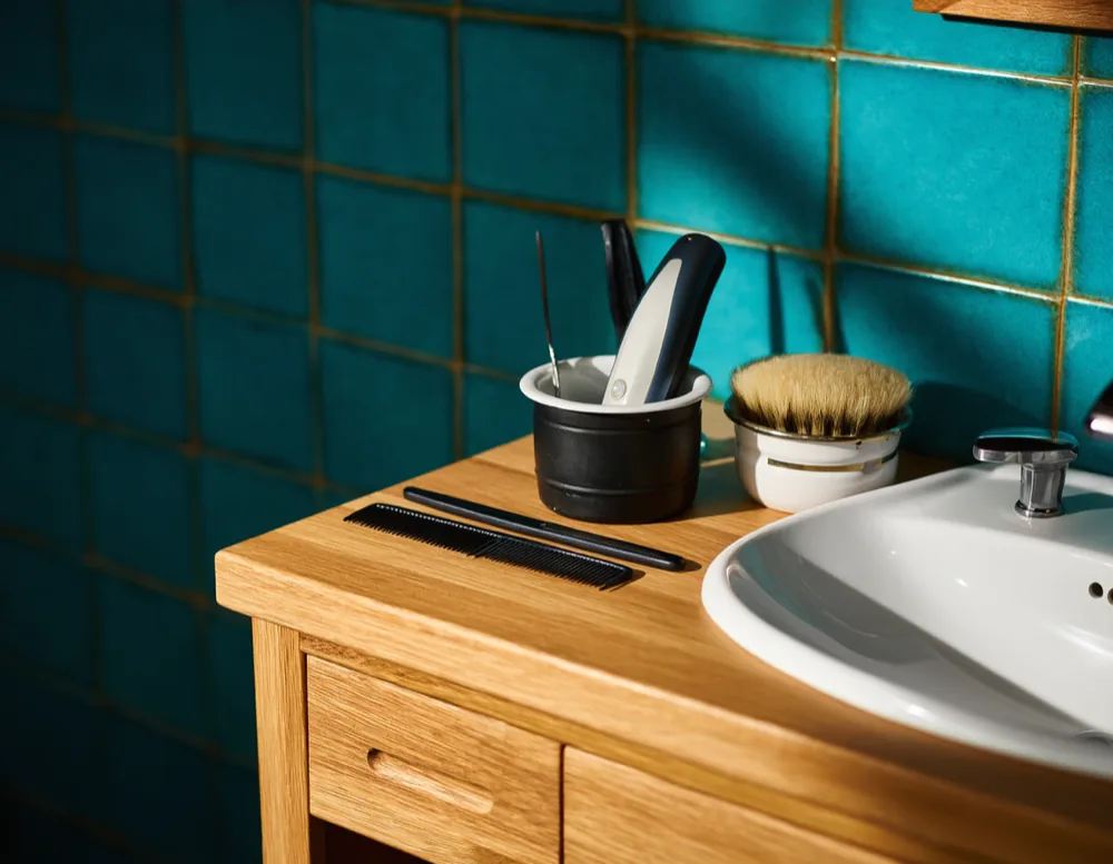 A straight razor, comb and shaving brush laid out on the oak counter in front of the teal tiled wall