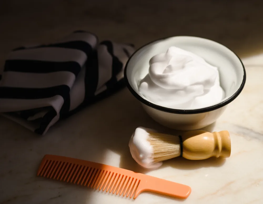 A bowl of shaving foam with a wooden brush beside a small folded barber cape